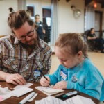 A girl in a blue sweater writes seated with a man in a plaid shirt.