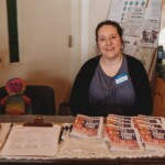 A woman sits at a table with sign in sheets and zines.
