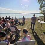 A presenter speaks to a seated group beside a lake.