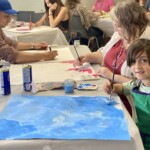 A group of children and adults sit at tables to paint.